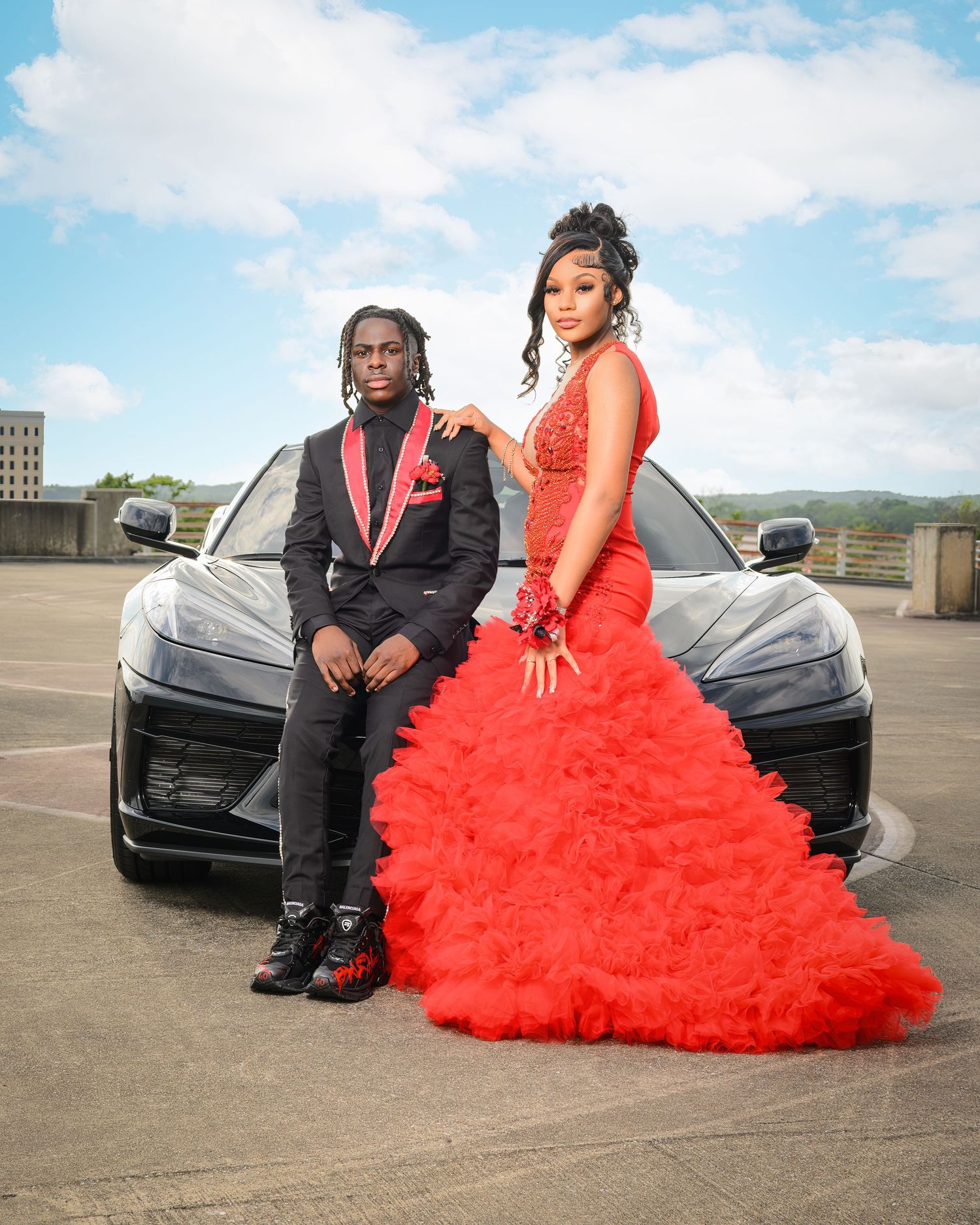 A couple poses in formal attire against a black car, with the woman wearing a dramatic red ruffled gown.