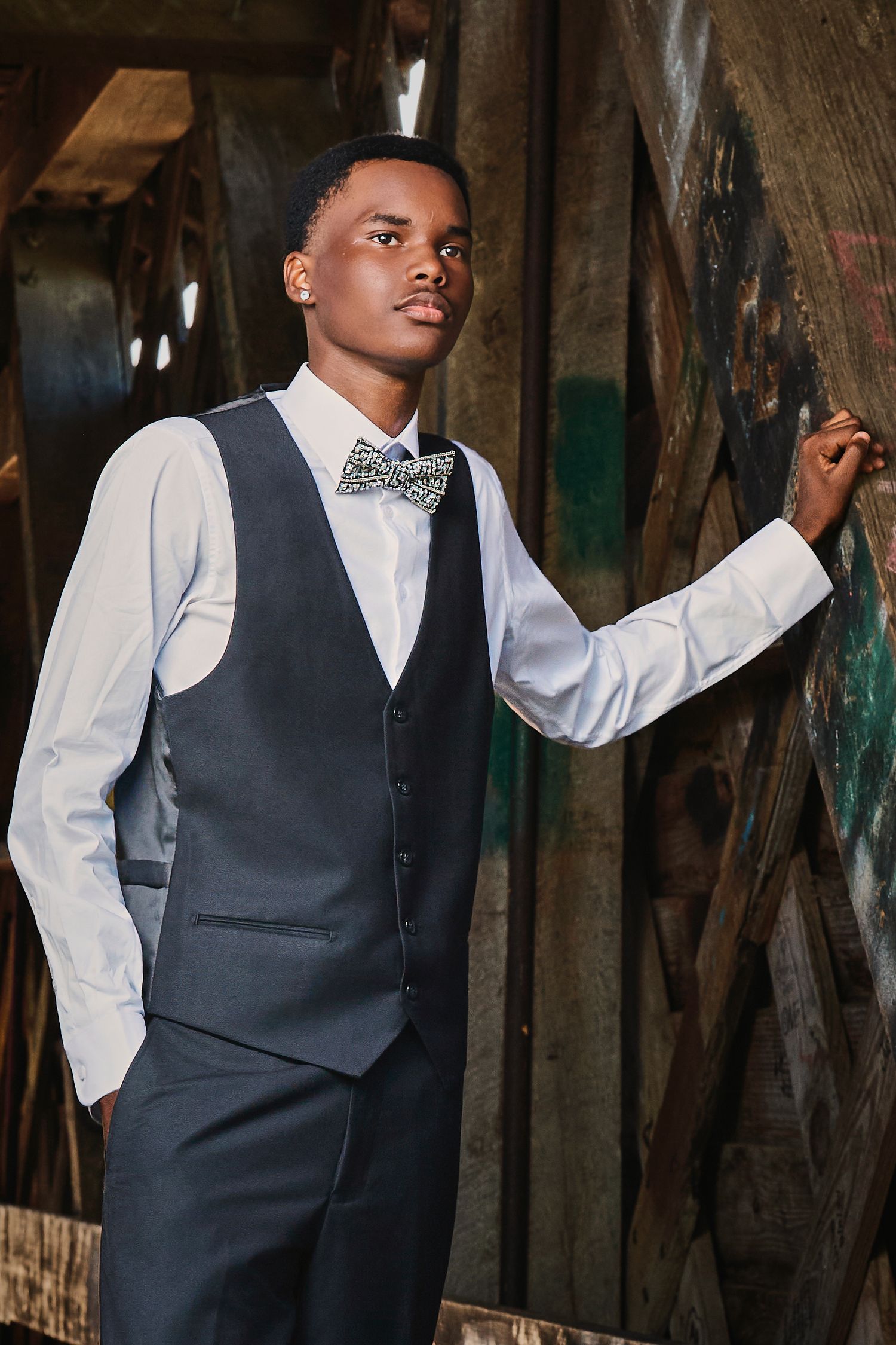 Young Black man in gray vest, white shirt, and bow tie poses confidently in an urban setting.