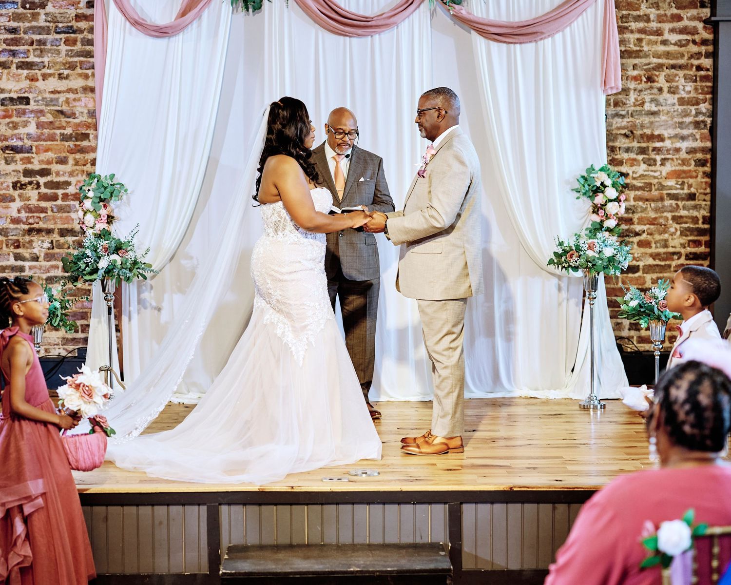 Couple during their wedding ceremony in front of draped fabric and brick wall.