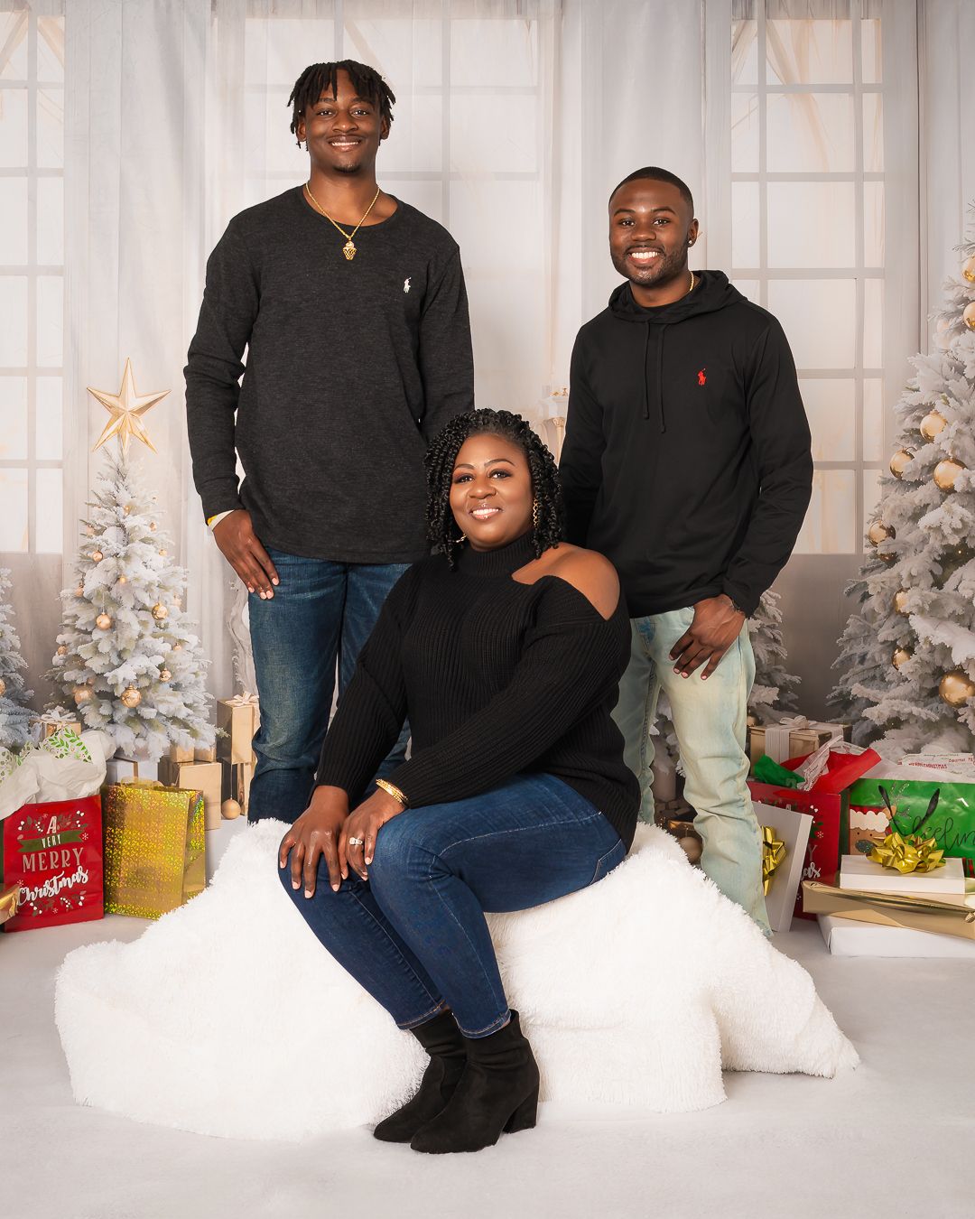 Three people in casual black sweaters pose together in a festive holiday setting with wrapped presents.