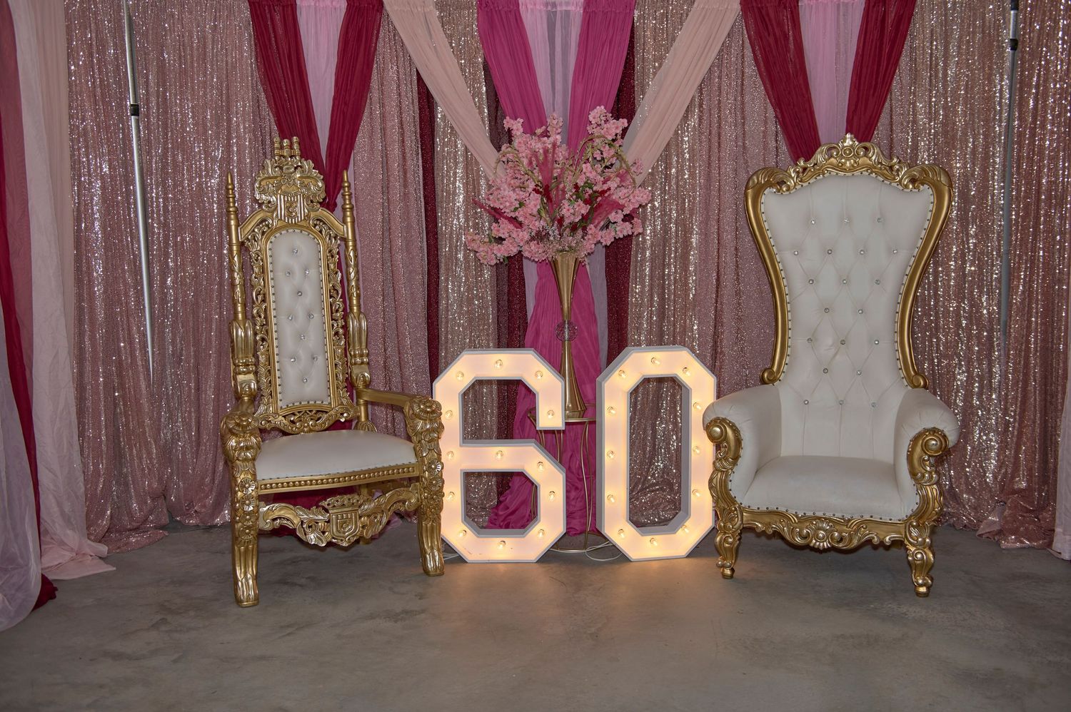 Elegant birthday party setup with illuminated number 60 between two ornate gold throne chairs against a pink draped backdrop.