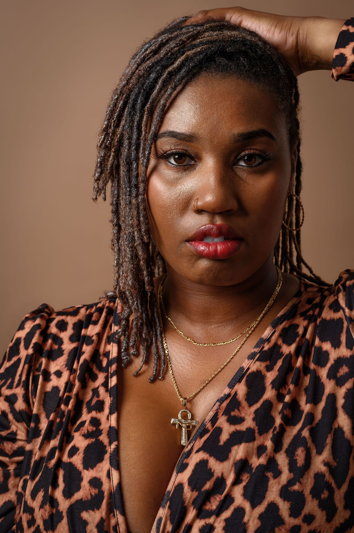 Woman with locs wearing leopard print top and gold cross necklace poses against a tan background.
