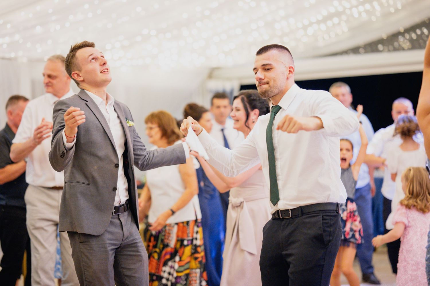 Wedding guests dance energetically at reception with bright lighting.
