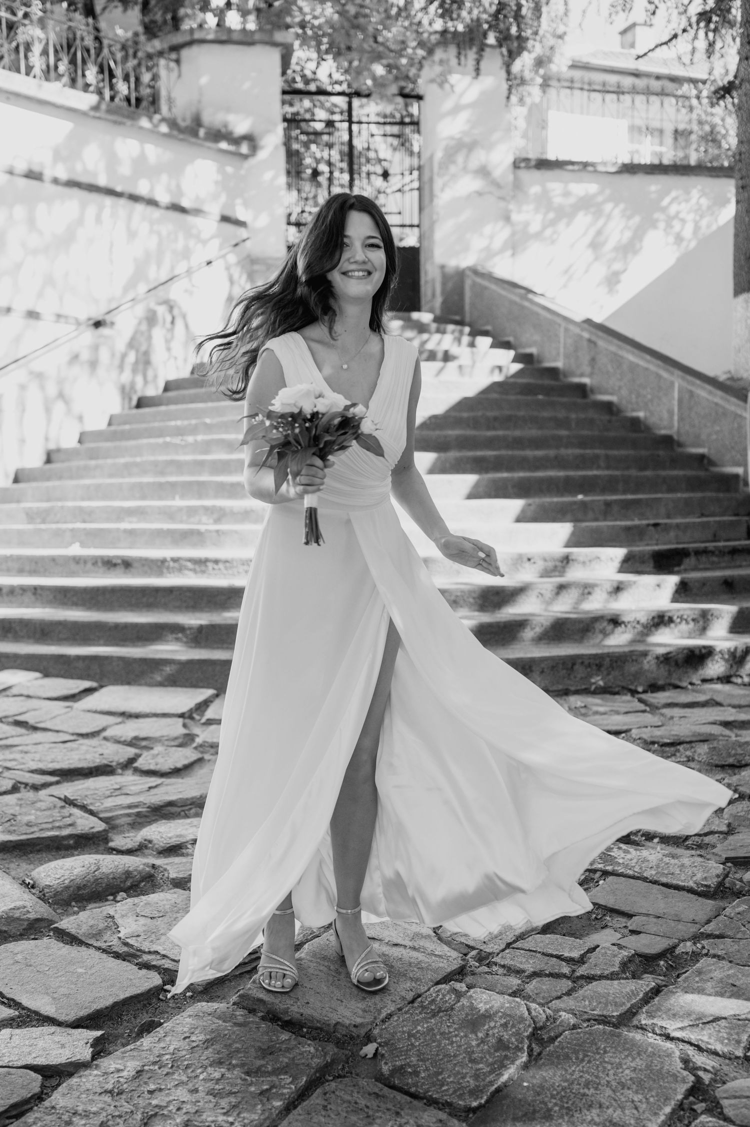 A flowing white wedding dress twirls on cobblestone steps in black and white photography.