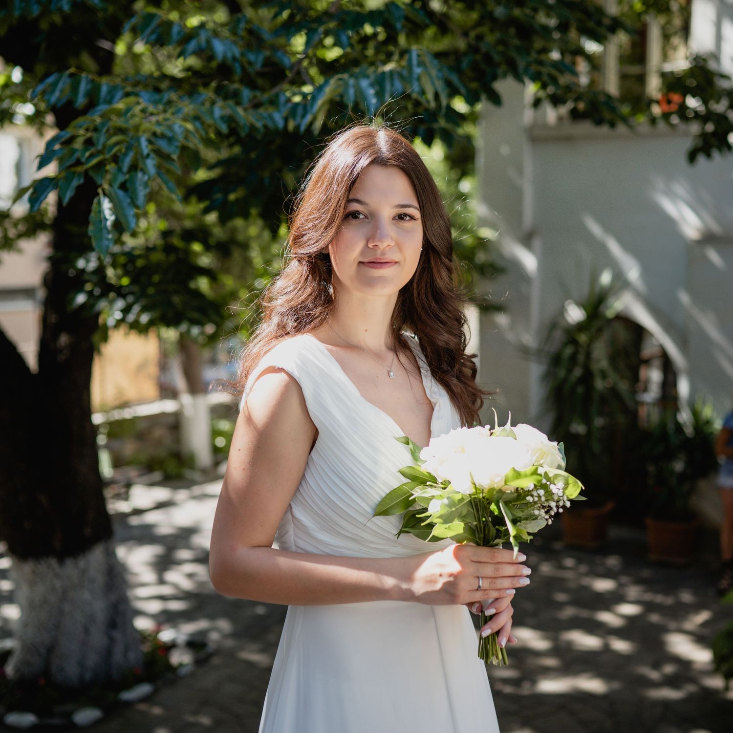 A bride in a white dress holds a small floral bouquet against a sunlit outdoor backdrop.