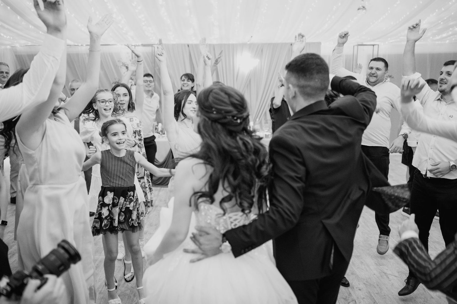 Wedding party dances energetically under white tent during reception celebration.