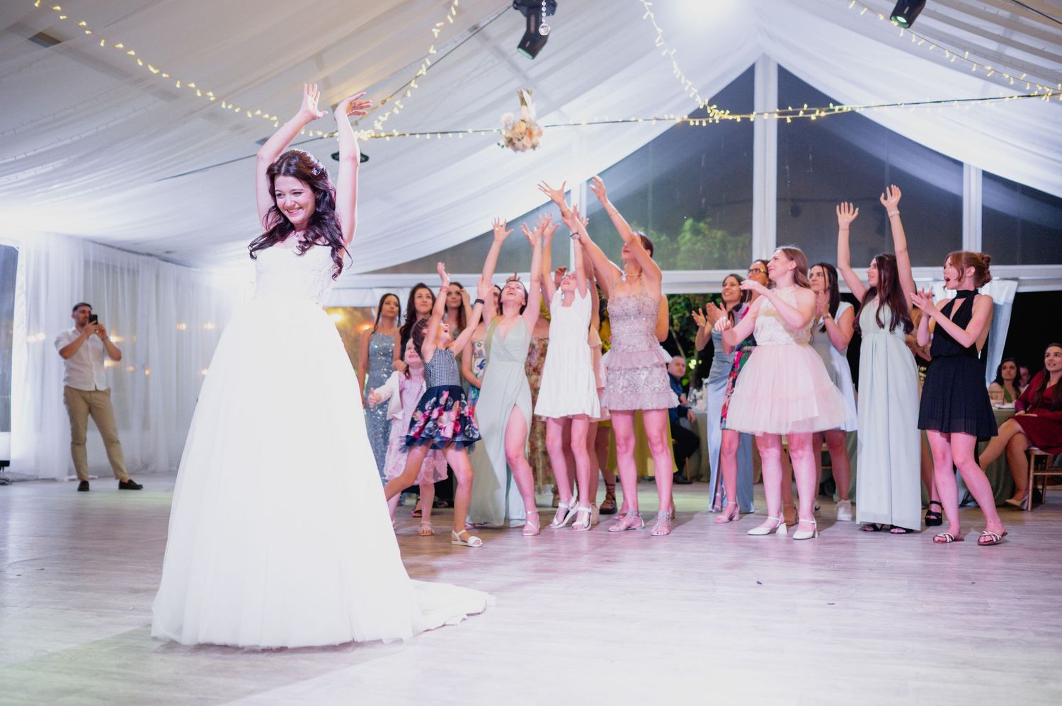 Wedding party guests gather on a dance floor with a classic tent canopy and string lights overhead.