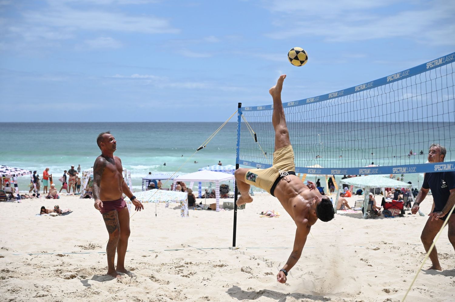 Man performs bicycle kick over volleyball net on sunny beach while onlookers watch in background.
