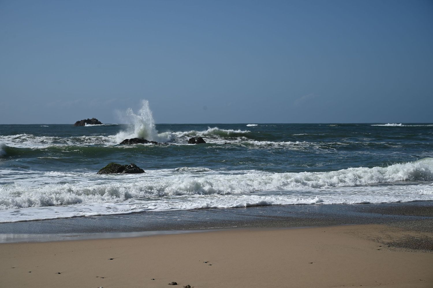 Waves crashing on rocks along a sandy beach shore with clear blue skies and open ocean in the background.