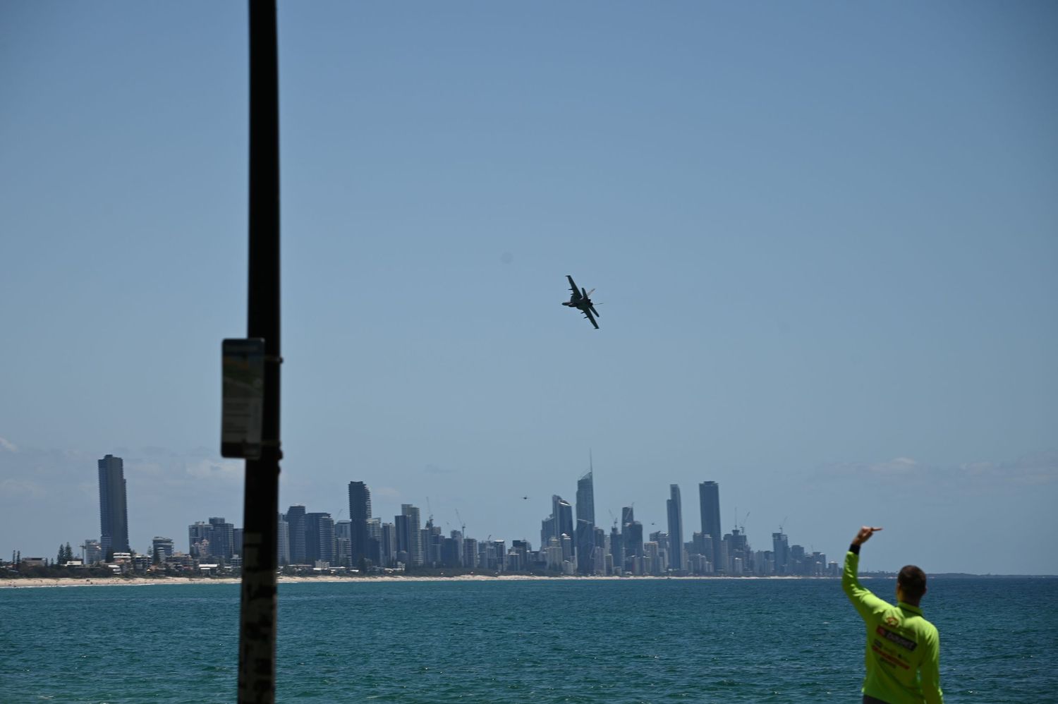 Person in yellow jacket watches jet fly over Gold Coast skyline from beach pier on clear sunny day.