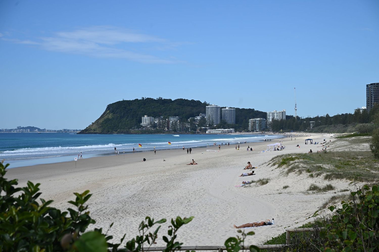 Panoramic view of a sandy beach with calm blue waters, lush headland, and coastal buildings under a clear sky.