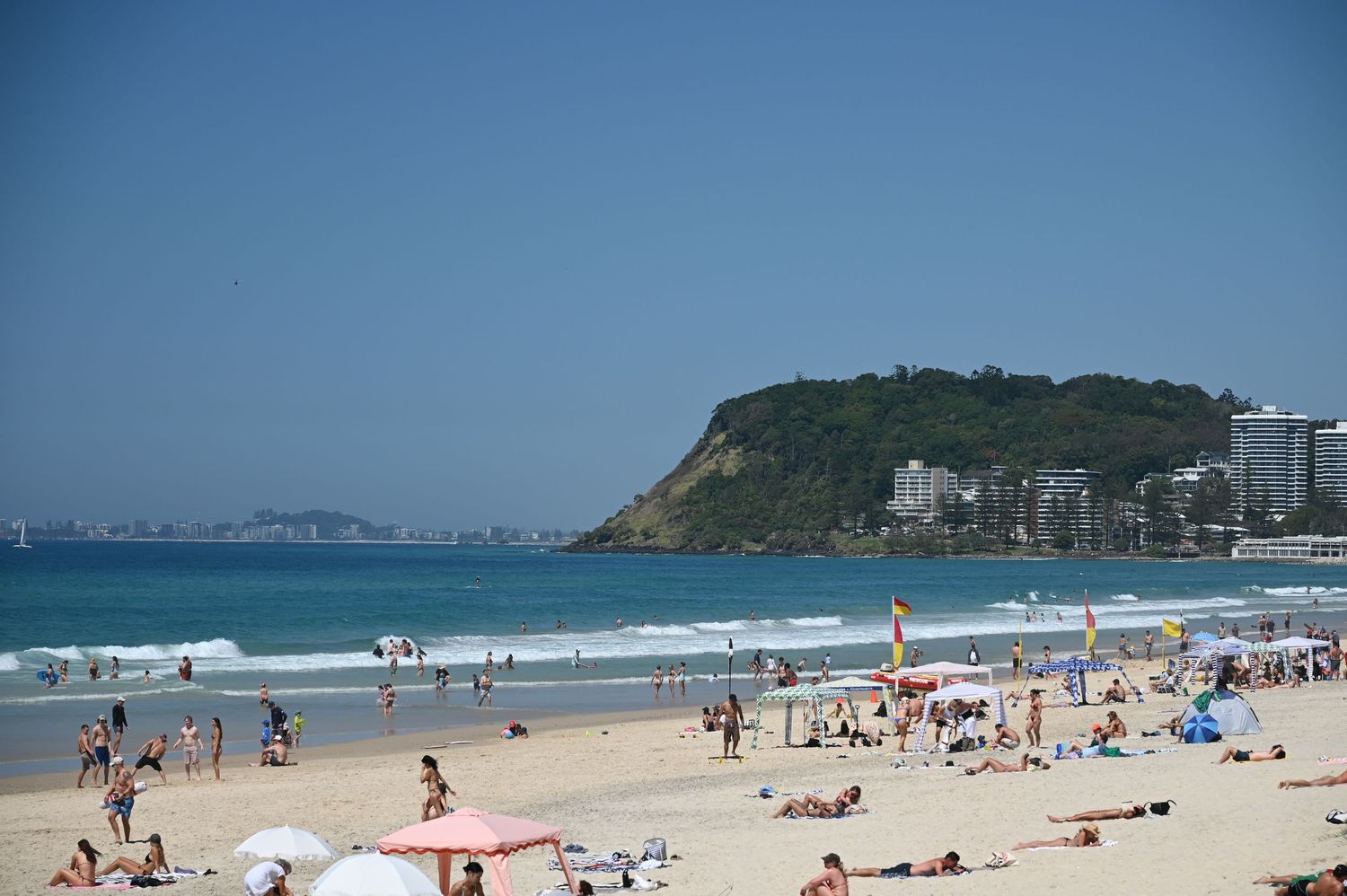 Crowded sandy beach with sunbathers, colorful umbrellas, calm blue ocean waves, and a green headland under clear blue skies.