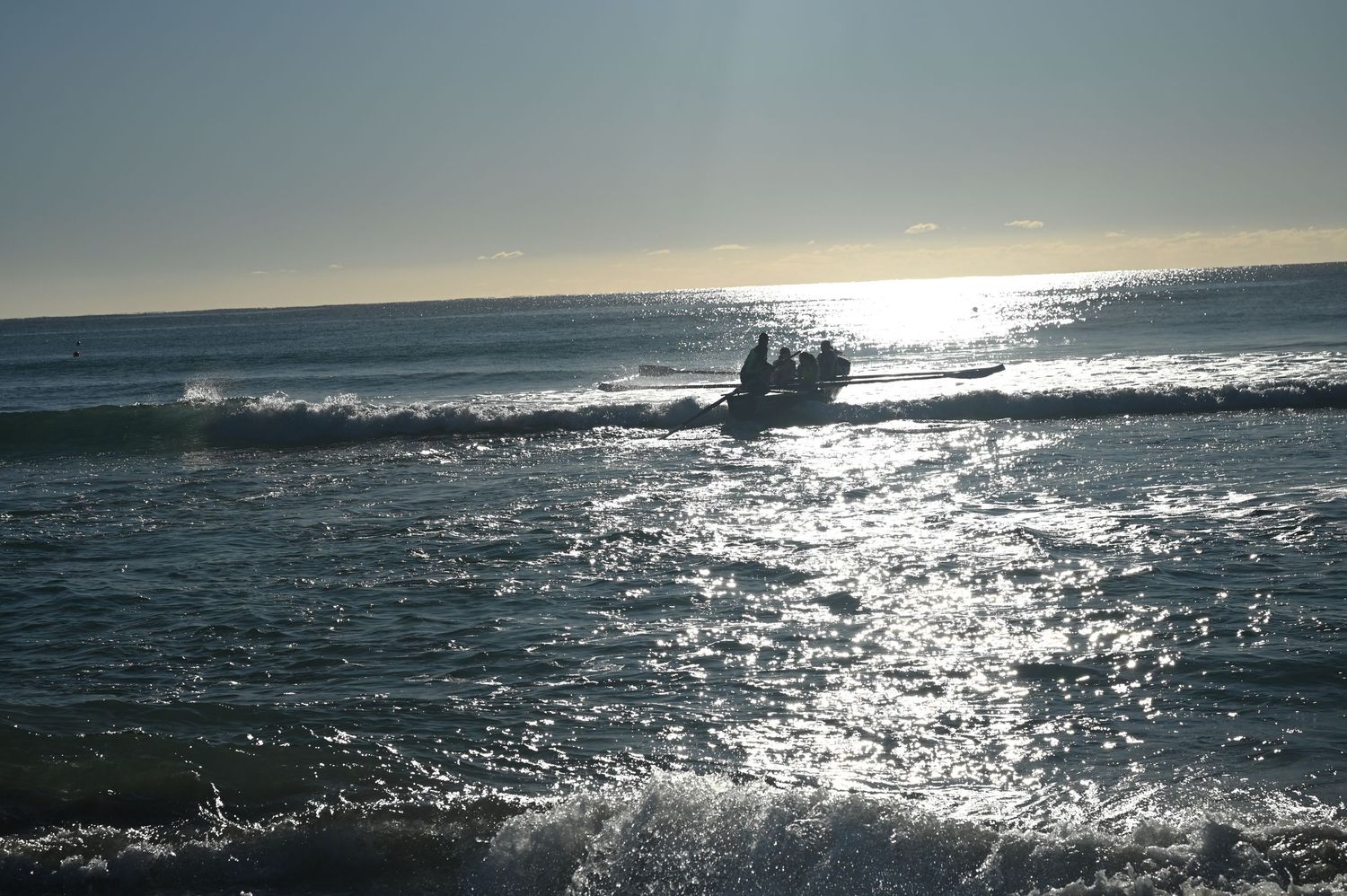 Small inflatable boat with passengers navigating through sparkling sunlit ocean waves near a sandy beach at sunset.