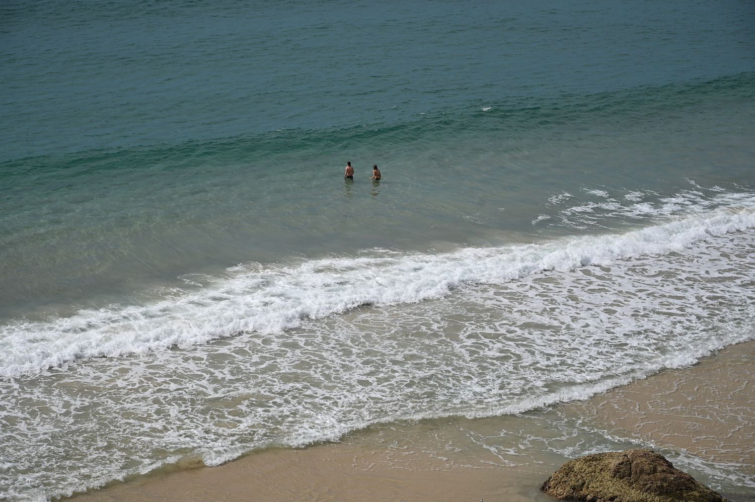 Two swimmers wade in clear turquoise ocean waves on a sandy beach, viewed from above.