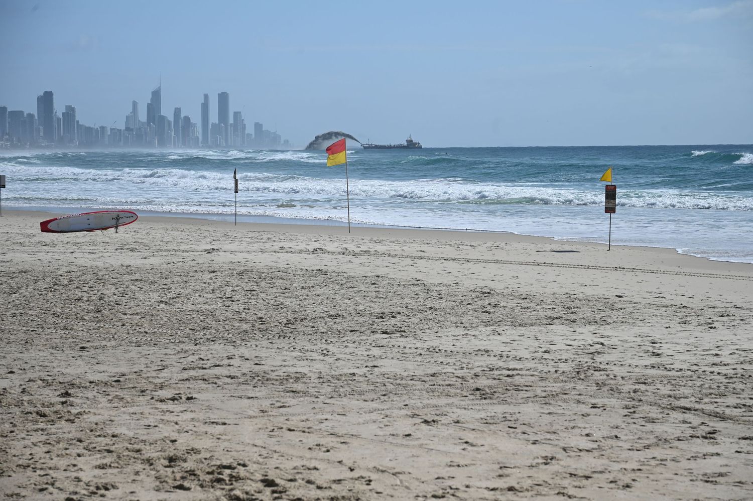 Sandy beach with a surfboard, colorful umbrella, and a city skyline visible across the ocean waves under a hazy sky.