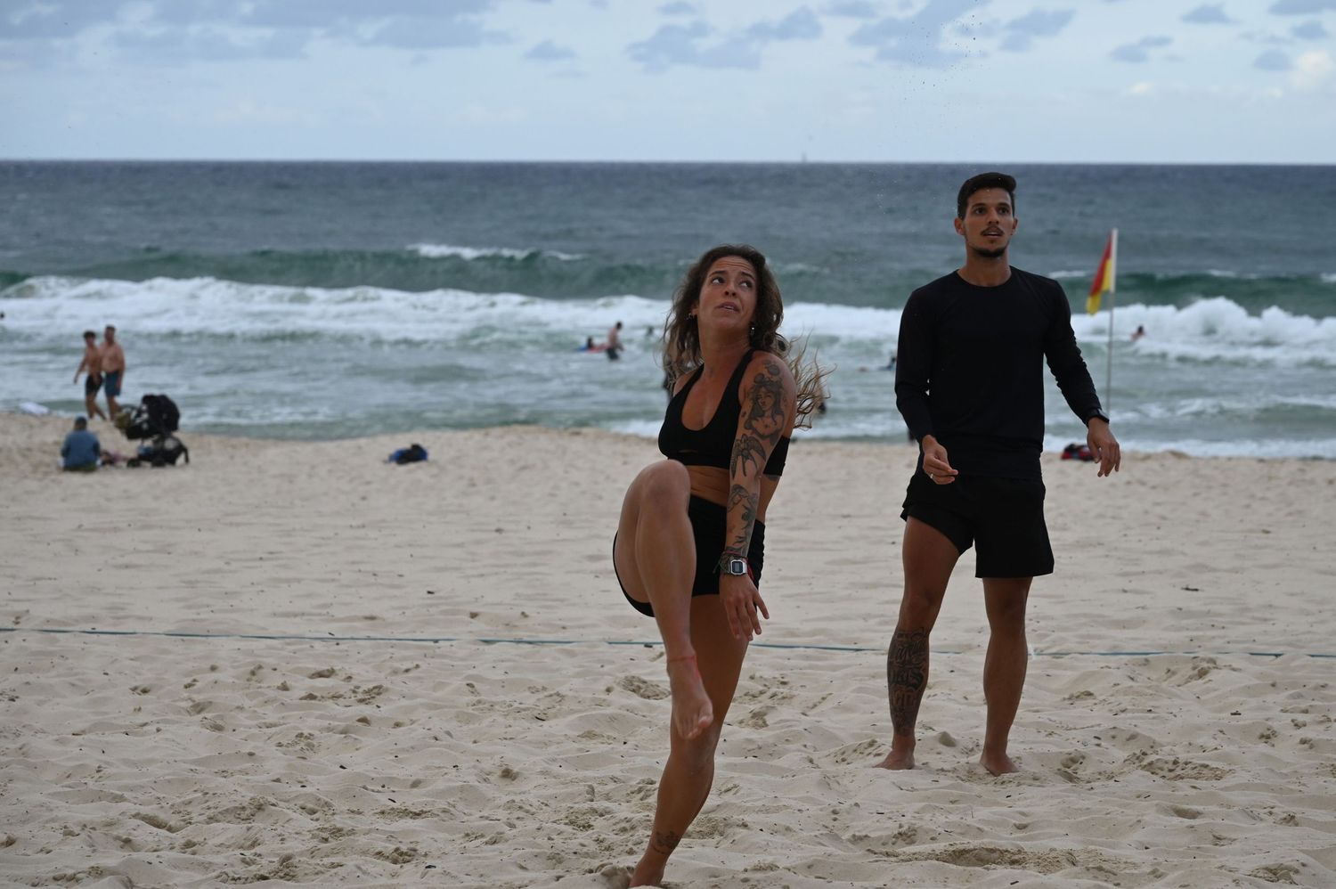 Tattooed woman and man play beach volleyball on sandy shore with ocean waves in background.