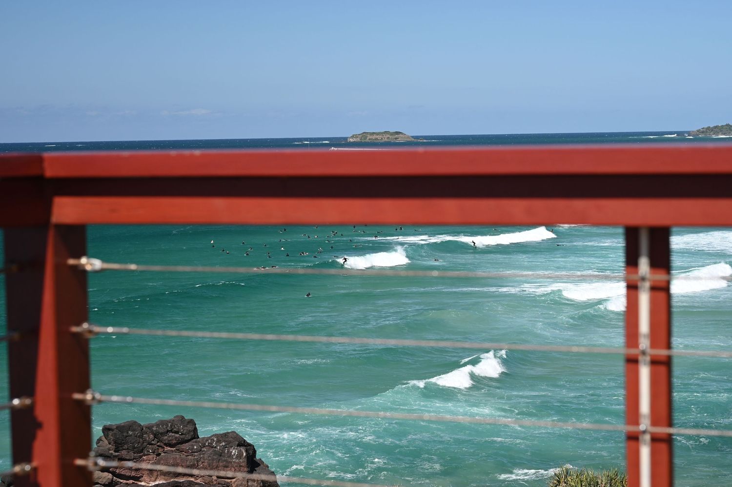 Turquoise ocean waves viewed through a red railing on a sunny day at a coastal lookout.