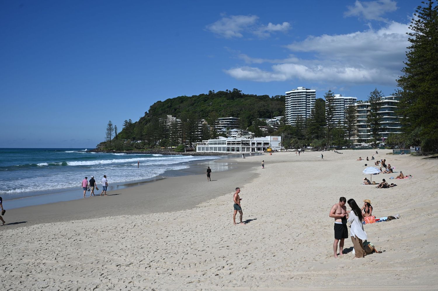 Sunny beach with white sand, blue ocean waves, green tree-covered headland, and high-rise buildings under a bright blue sky.