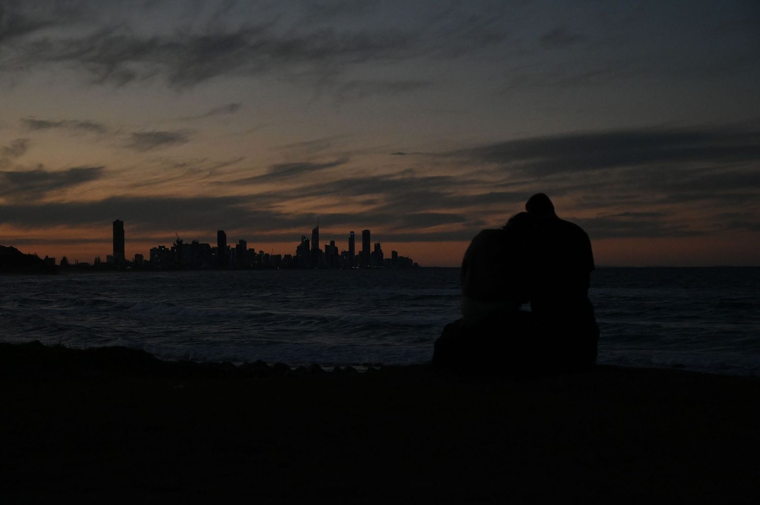 Silhouette of a couple sitting together on a beach at sunset with a city skyline in the background.