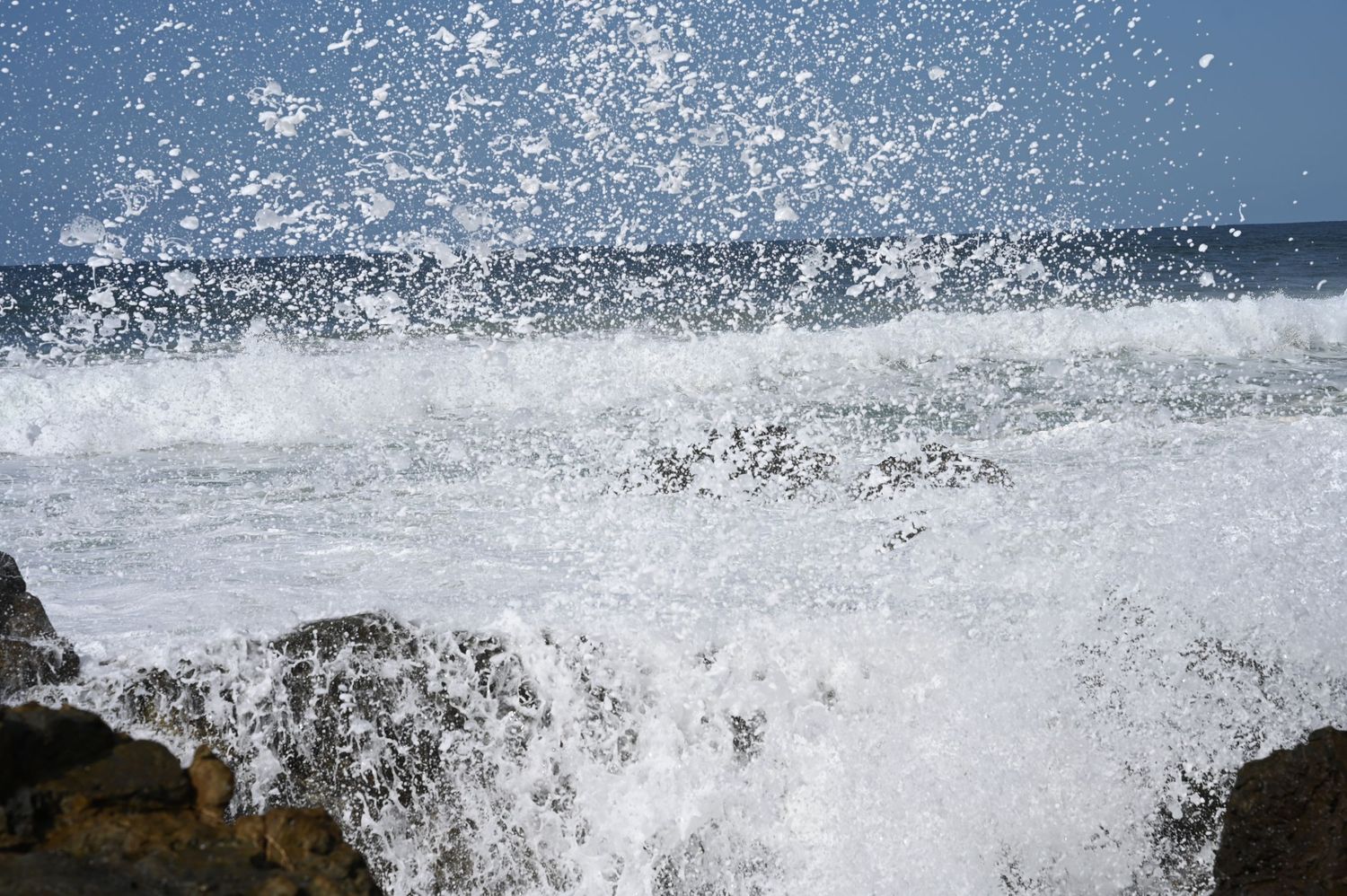 Powerful ocean waves crashing against rocky shoreline with white sea spray and foam under a clear blue sky.