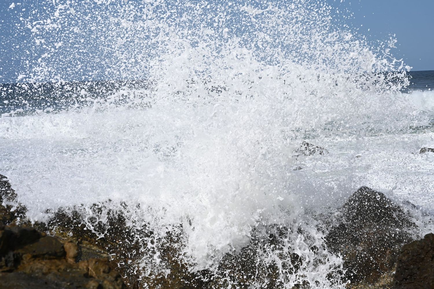 Powerful ocean waves crashing dramatically against rocky shoreline, creating white sea foam spray against a blue sky.