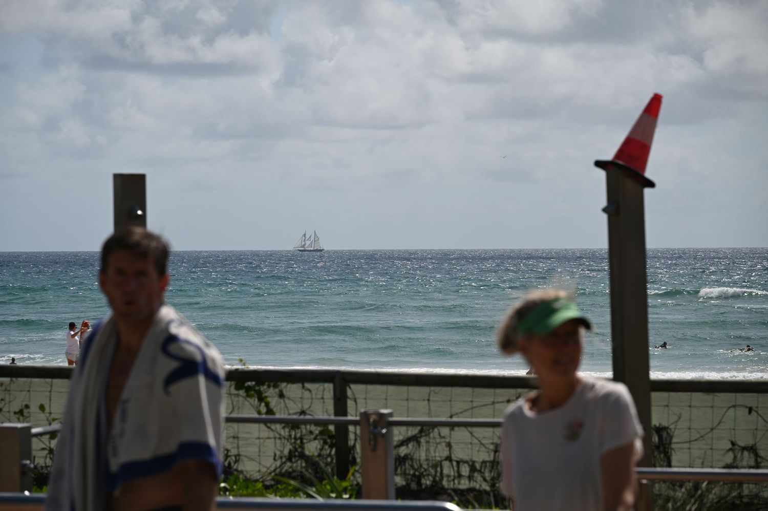 Two people walk near a beachside railing with ocean waves and a ship visible in the background under cloudy skies.