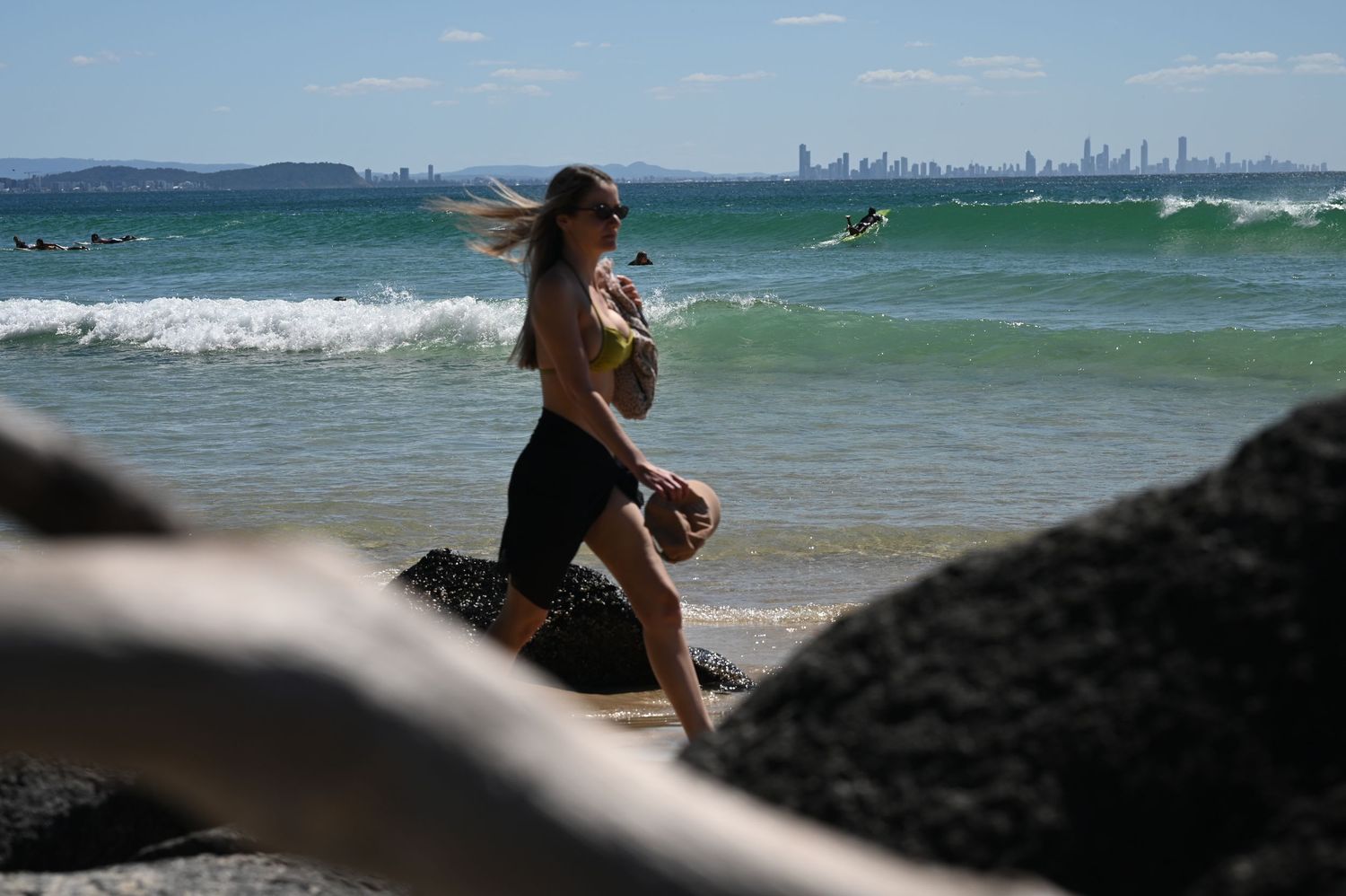 Woman walking on rocky beach with turquoise waves and city skyline in background on sunny day.