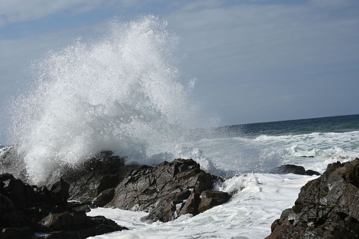 Powerful ocean wave crashes dramatically over dark coastal rocks under cloudy sky.