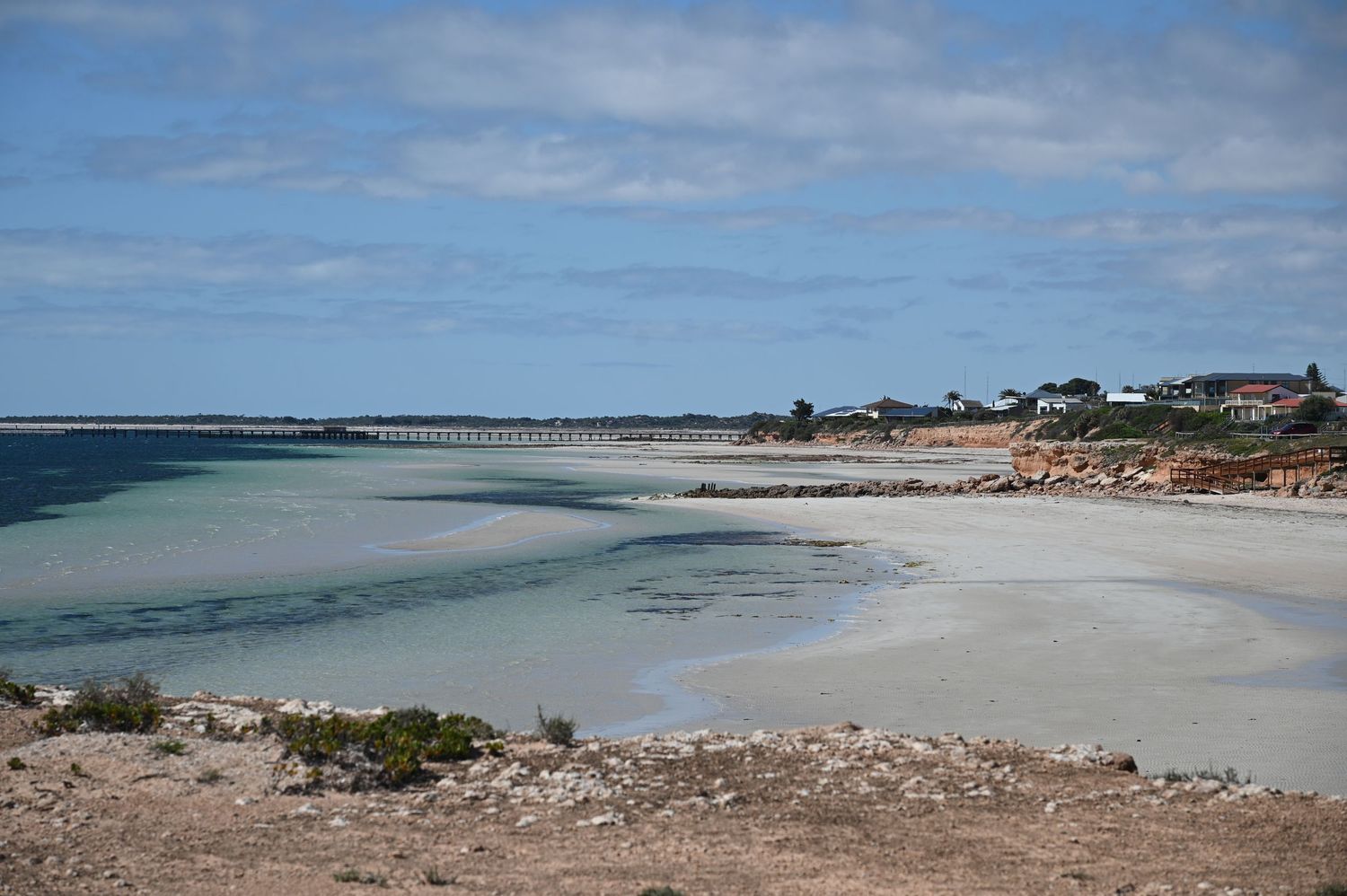 Serene Australian beach with white sand, turquoise water, rocky shoreline, and a distant pier under a partly cloudy sky.