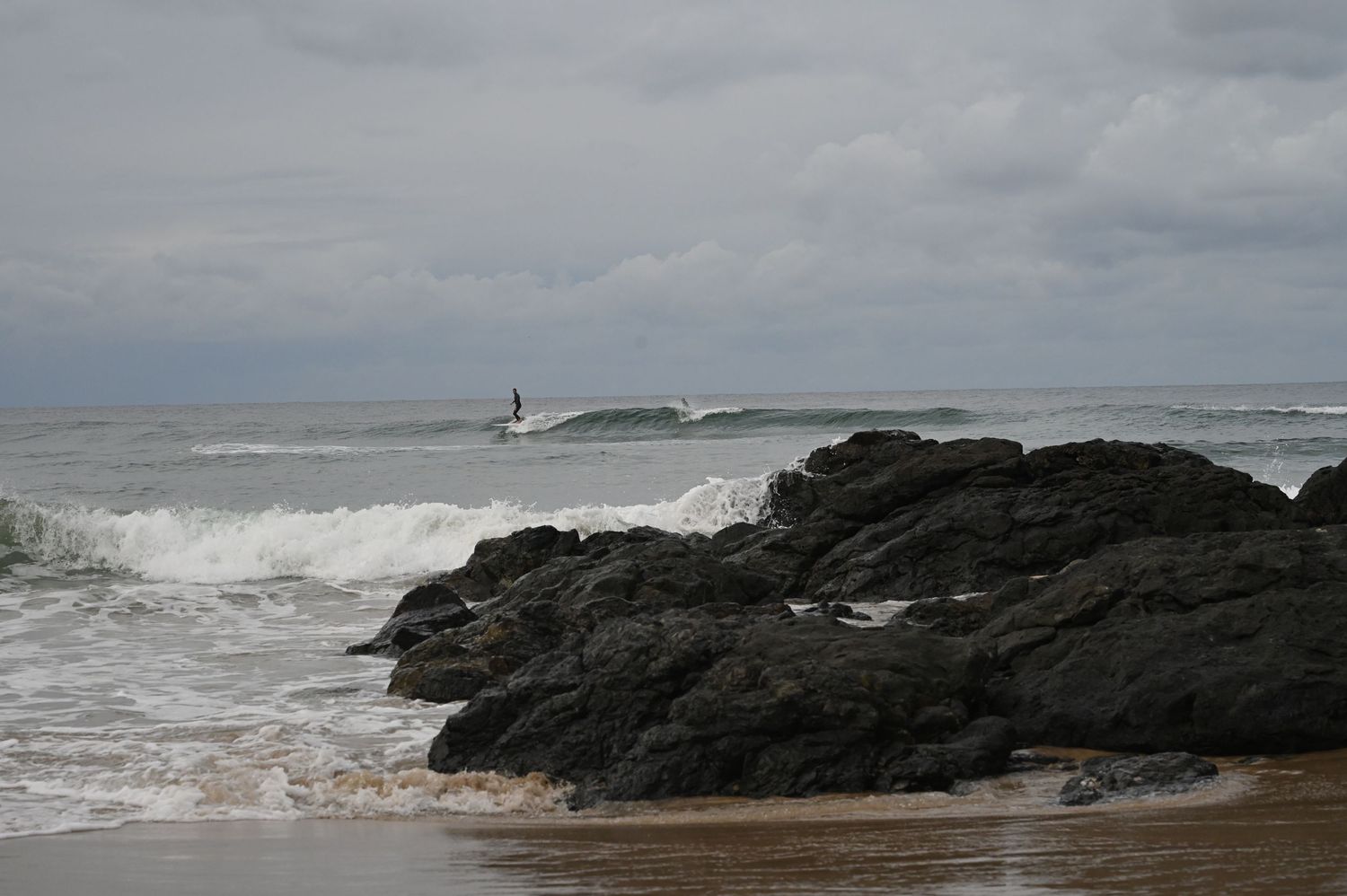 Rocky shoreline with crashing waves and a surfer riding in the distance under a cloudy sky.