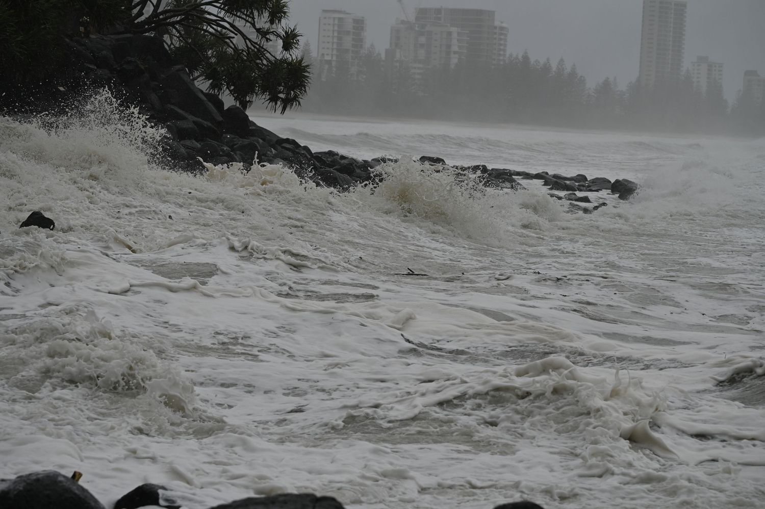 Turbulent stormy ocean waves battering a rocky shoreline with city buildings visible in the background.