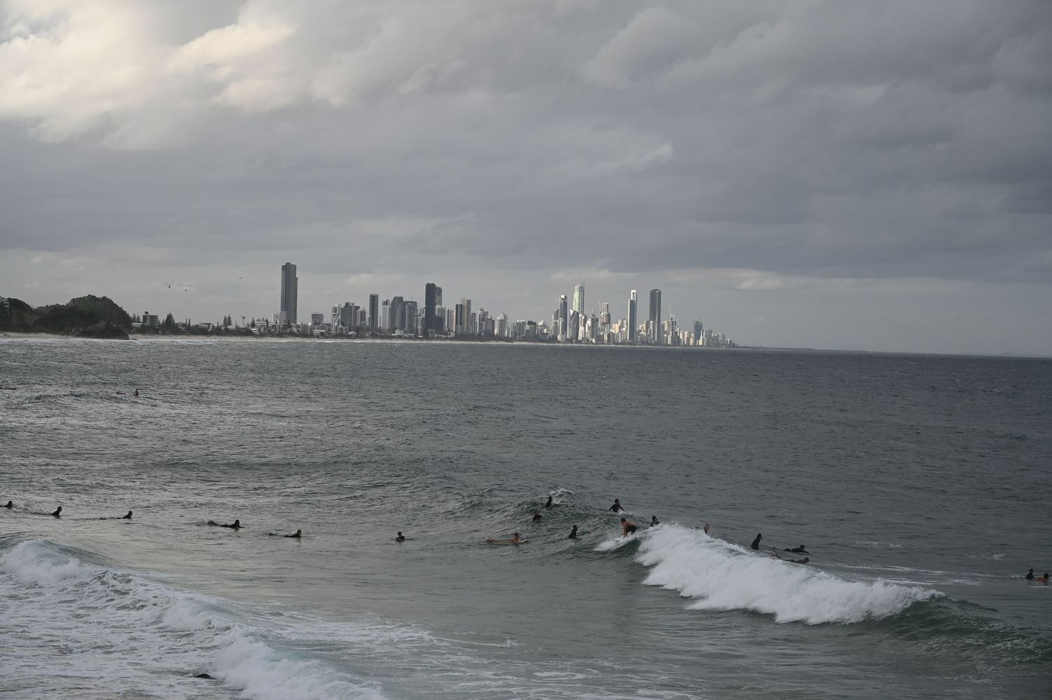 Surfers wait for waves in ocean with stunning Gold Coast city skyline under dramatic cloudy sky.