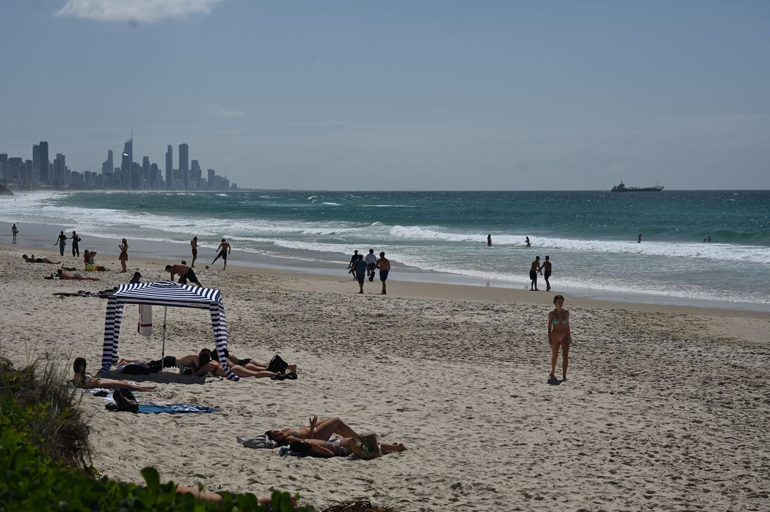 Beachgoers relax on a sunny Gold Coast beach with city skyline and ocean waves in the background.
