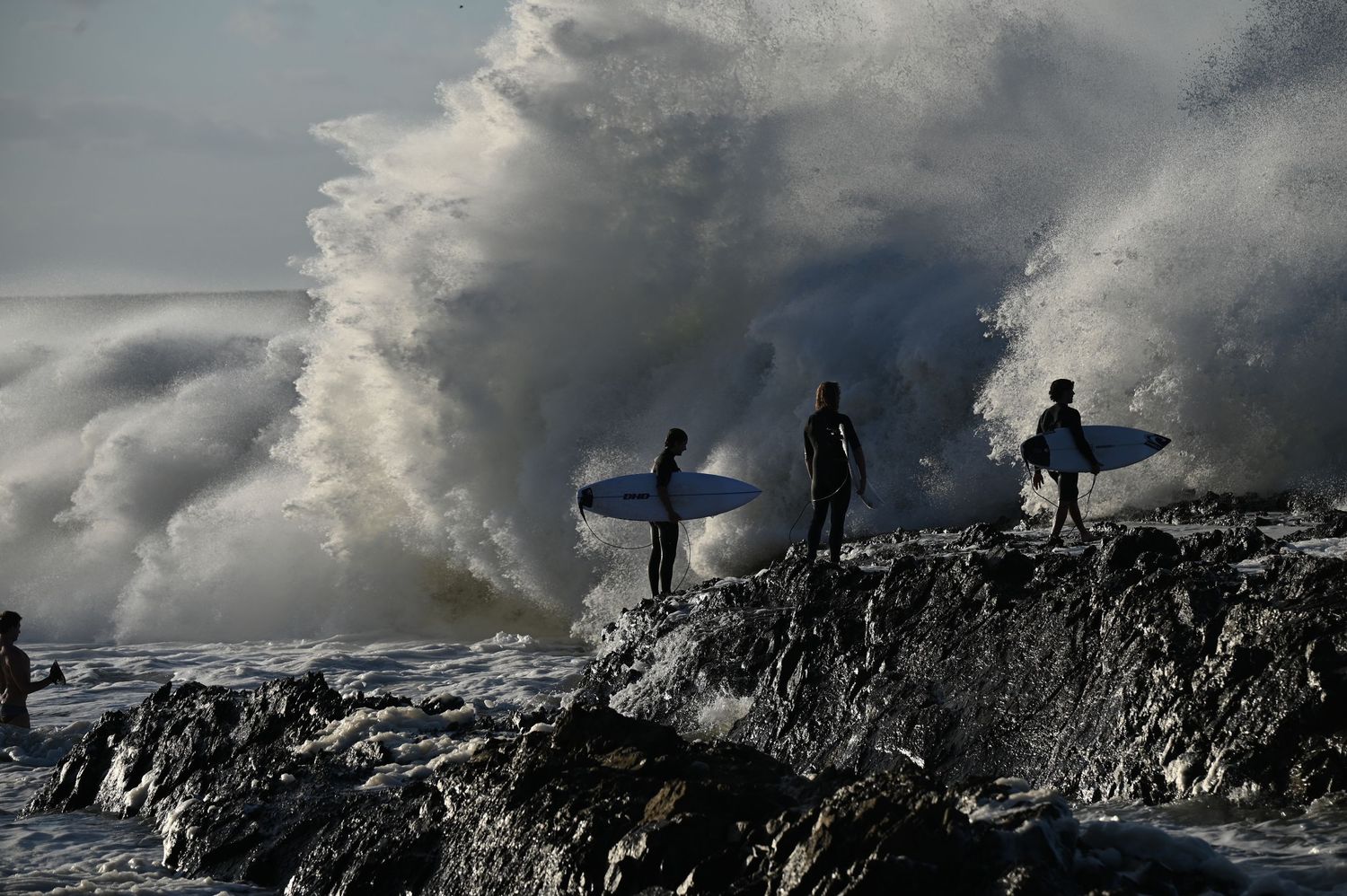 Brave surfers stand on rocky shore as massive powerful wave crashes dramatically behind them.