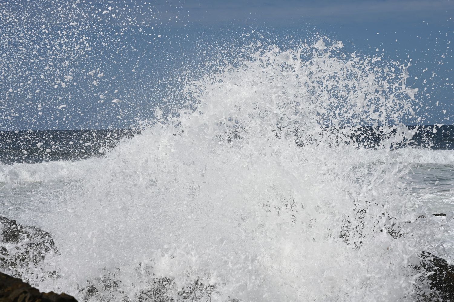 Powerful ocean wave crashing against rocky shore, sending white sea spray into blue sky.