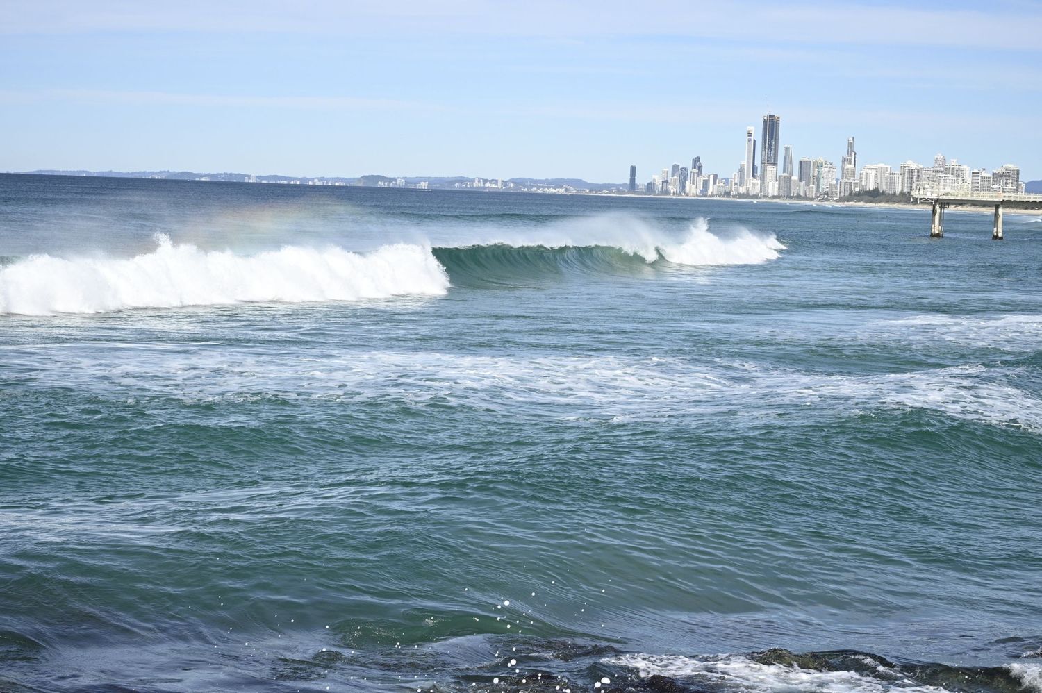 Surfer riding a wave at Gold Coast beach with city skyline visible in the background on a sunny day.