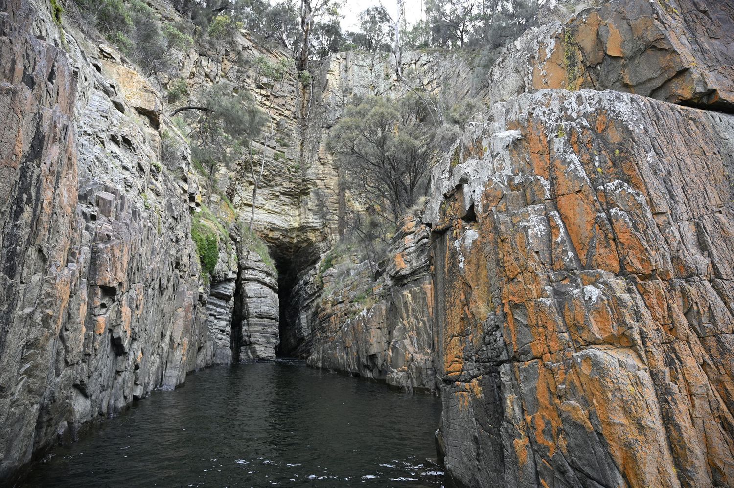 Narrow rocky gorge with dark water flowing through towering orange and grey cliff walls with a cave opening visible.