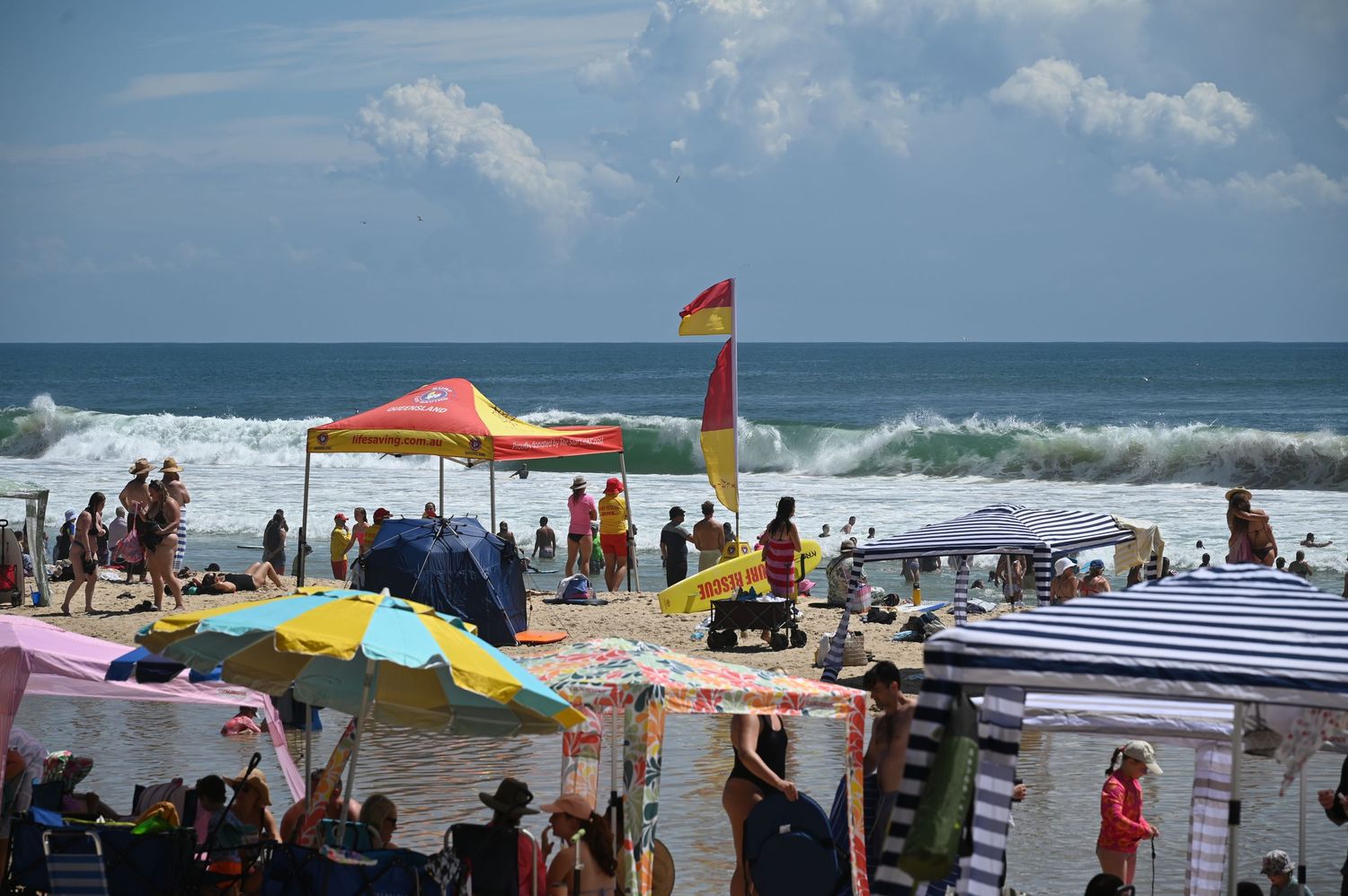 Crowded beach scene with colorful umbrellas, tents, and a lifeguard flag amid crashing ocean waves on a sunny day.