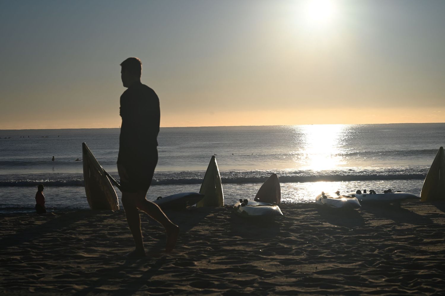 Silhouette of a man standing on a sandy beach at sunset with surfboards and ocean in the background.