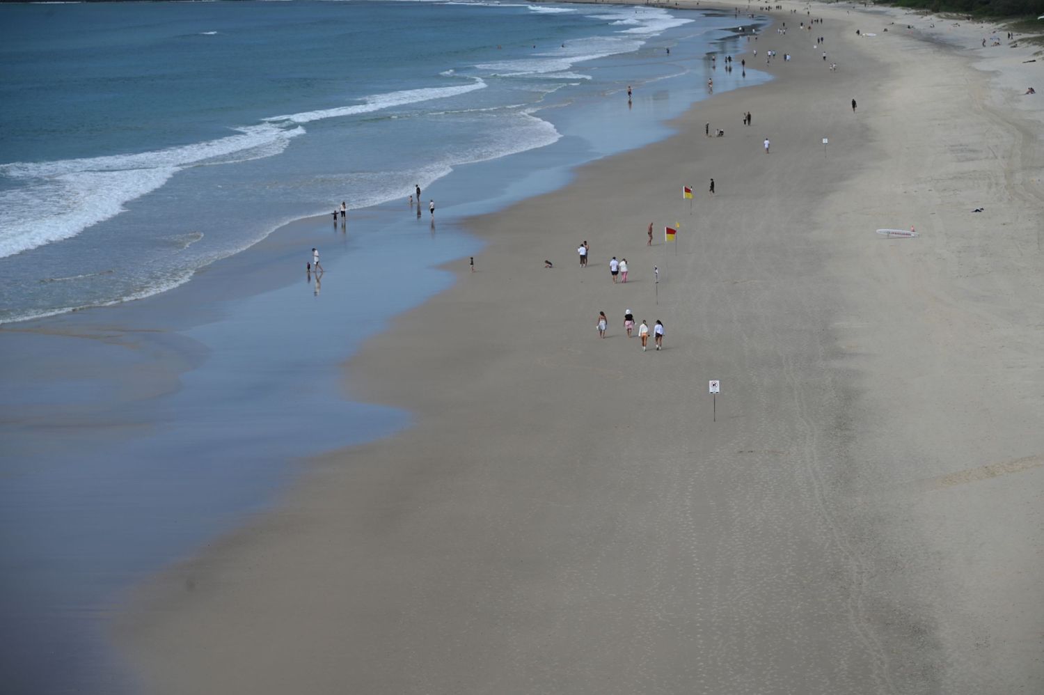 Aerial view of a sandy beach with gentle waves and scattered people walking along the shoreline on a cloudy day.