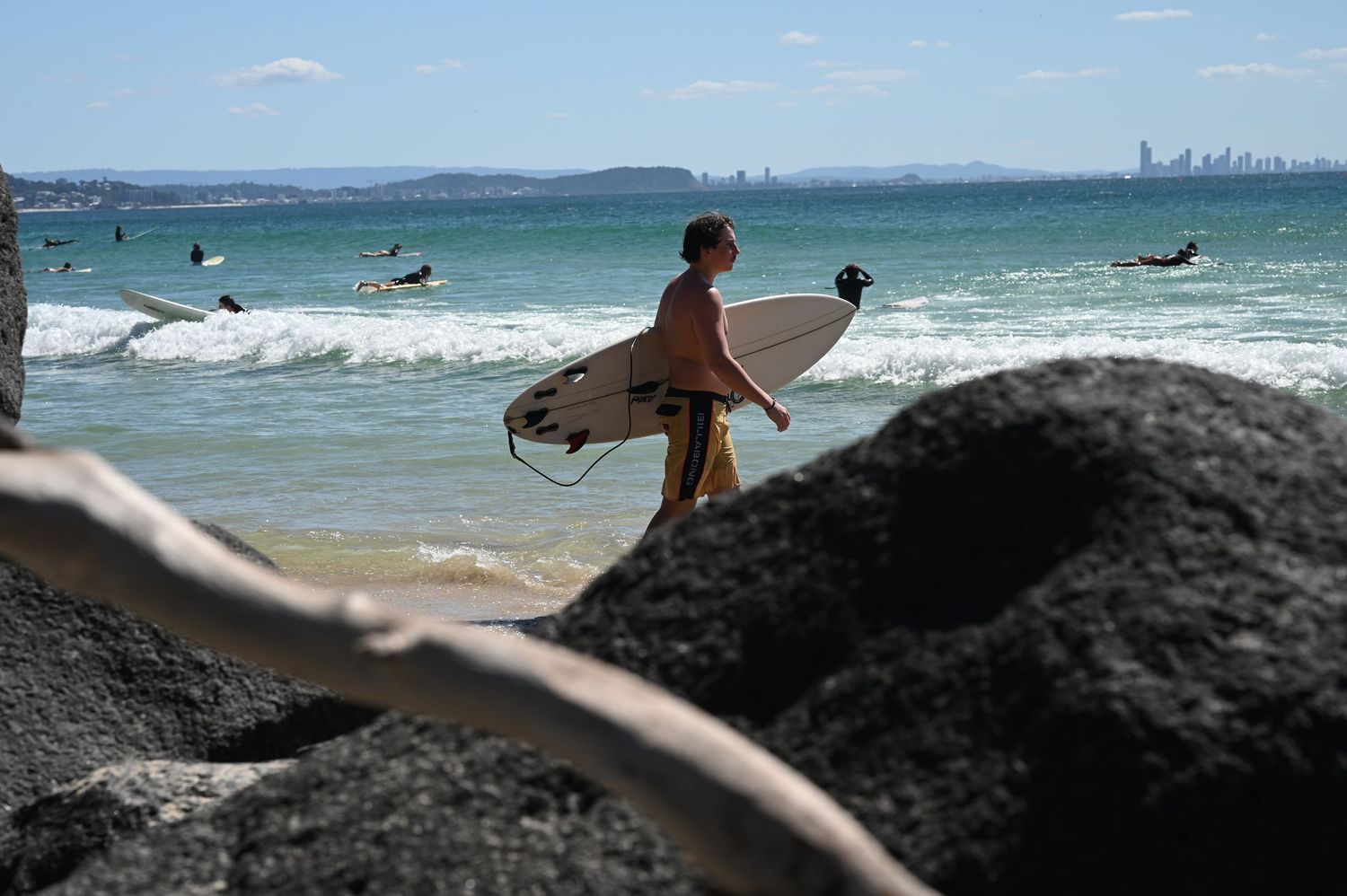 Surfer carrying a white surfboard walks along a rocky beach with ocean waves and city skyline in the background.