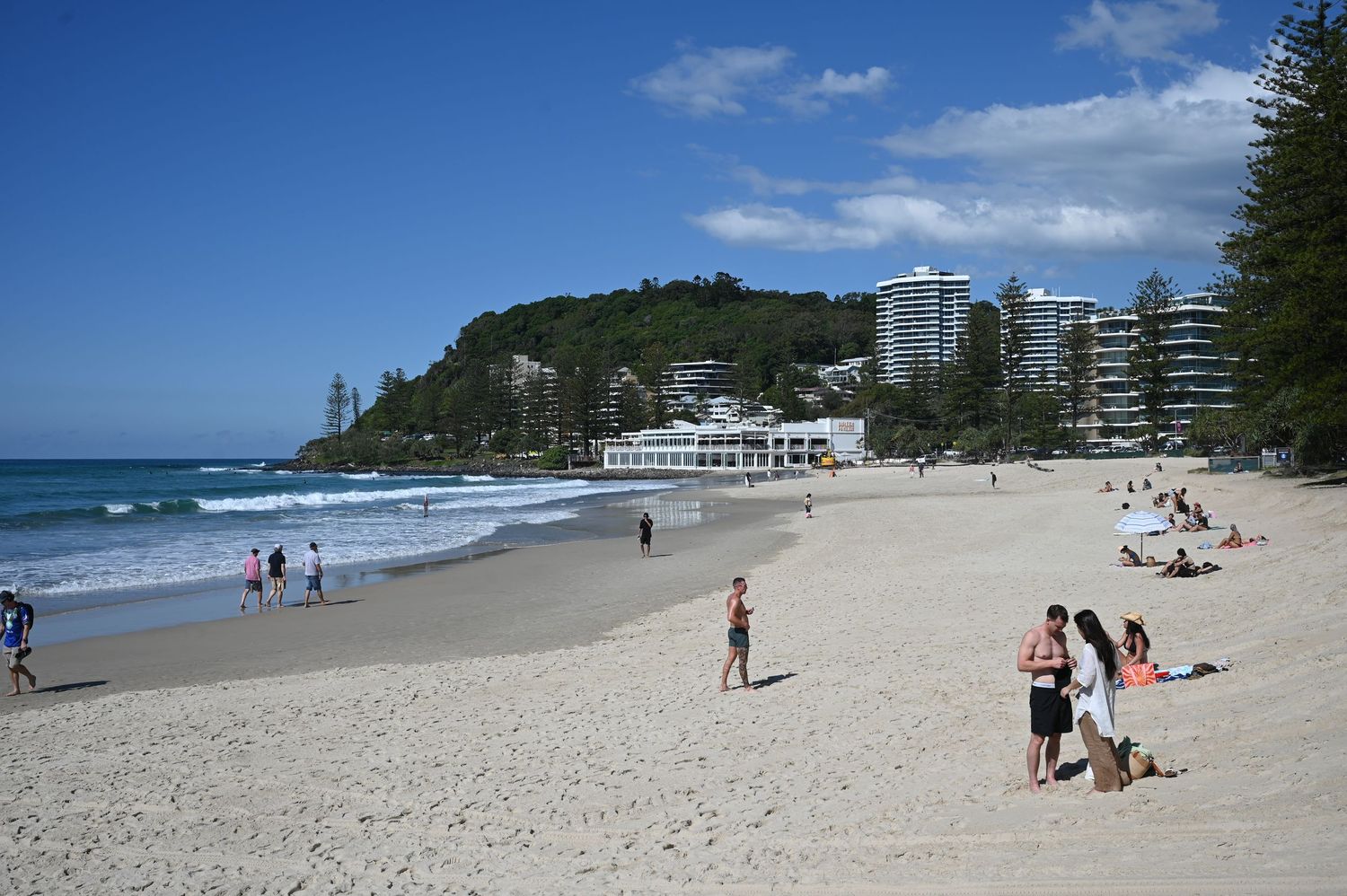 Sunny beach with white sand, blue waves, tall pine trees, and modern buildings on a hillside under a bright blue sky.