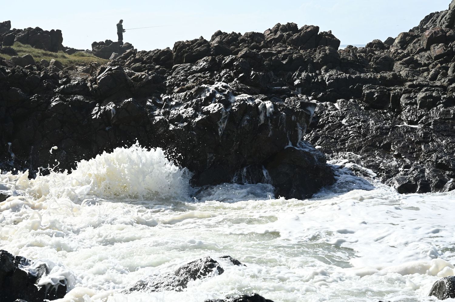 Powerful ocean waves crashing against dark volcanic rocks on a rugged coastline, with a person standing on the cliff above.