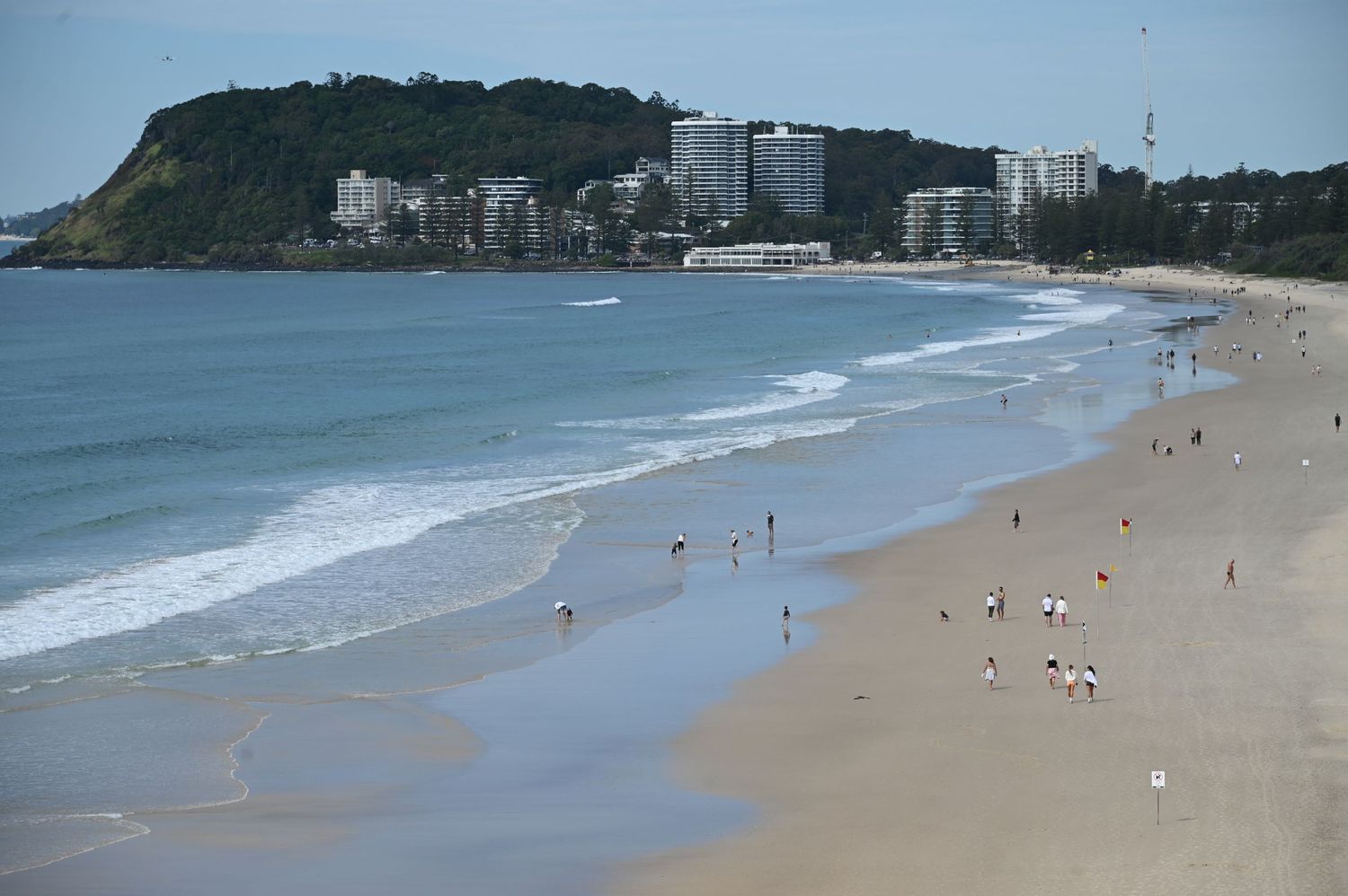 Aerial view of a sandy beach with gentle waves, scattered beachgoers, and coastal buildings on a green hillside.