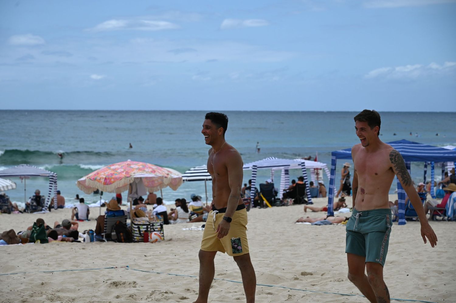 Two shirtless men walking on a sunny beach with colorful umbrellas and ocean waves in the background.