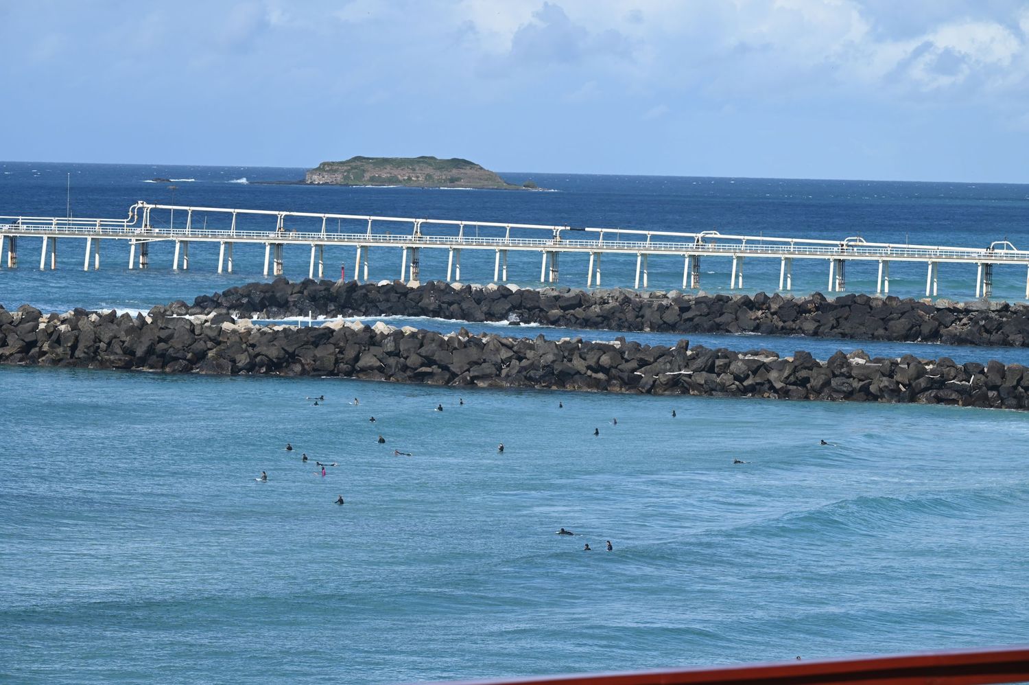 Aerial view of a coastal bridge over turquoise waters with rocky breakwater and small tropical island in background.