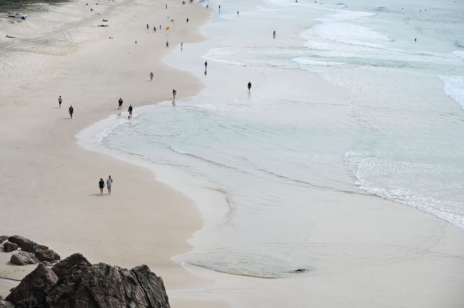 Aerial view of people walking on a wide sandy beach with gentle ocean waves and rocky shoreline.