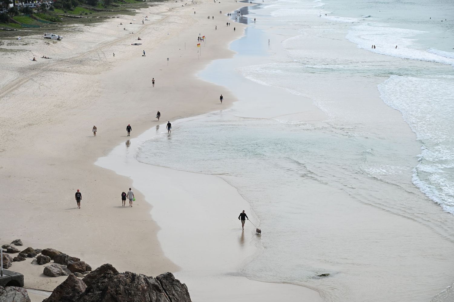 People walking along a wide sandy beach with gentle waves on a cloudy day, viewed from elevated rocky vantage point.
