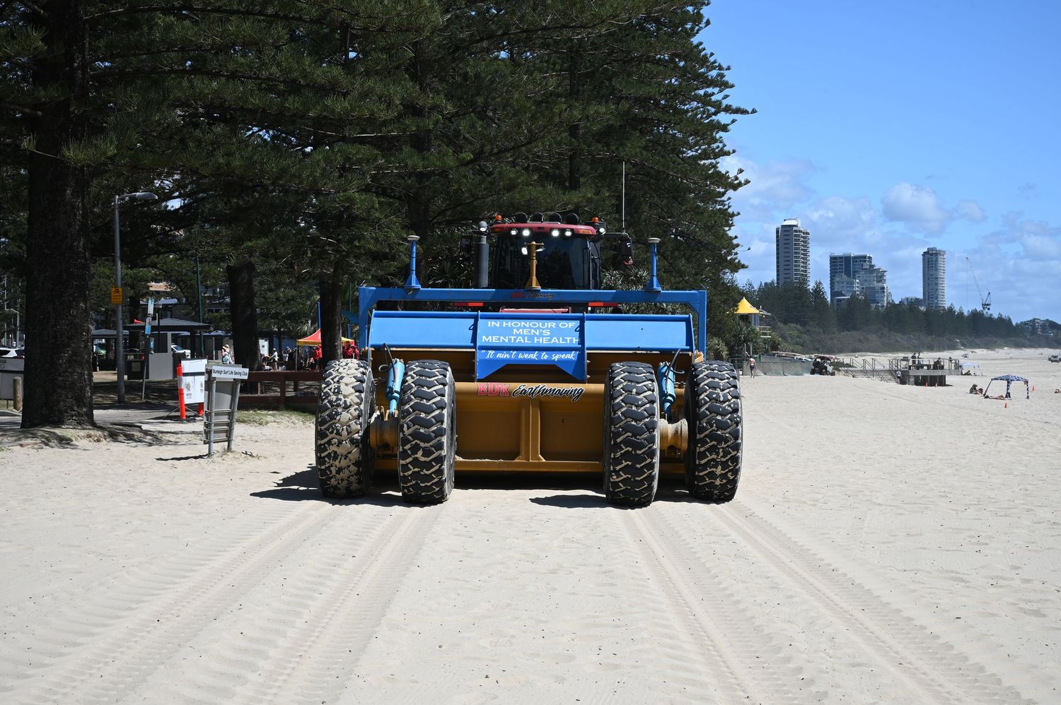 Blue beach cleaning tractor on sandy Gold Coast beach with city skyline and pine trees in background.