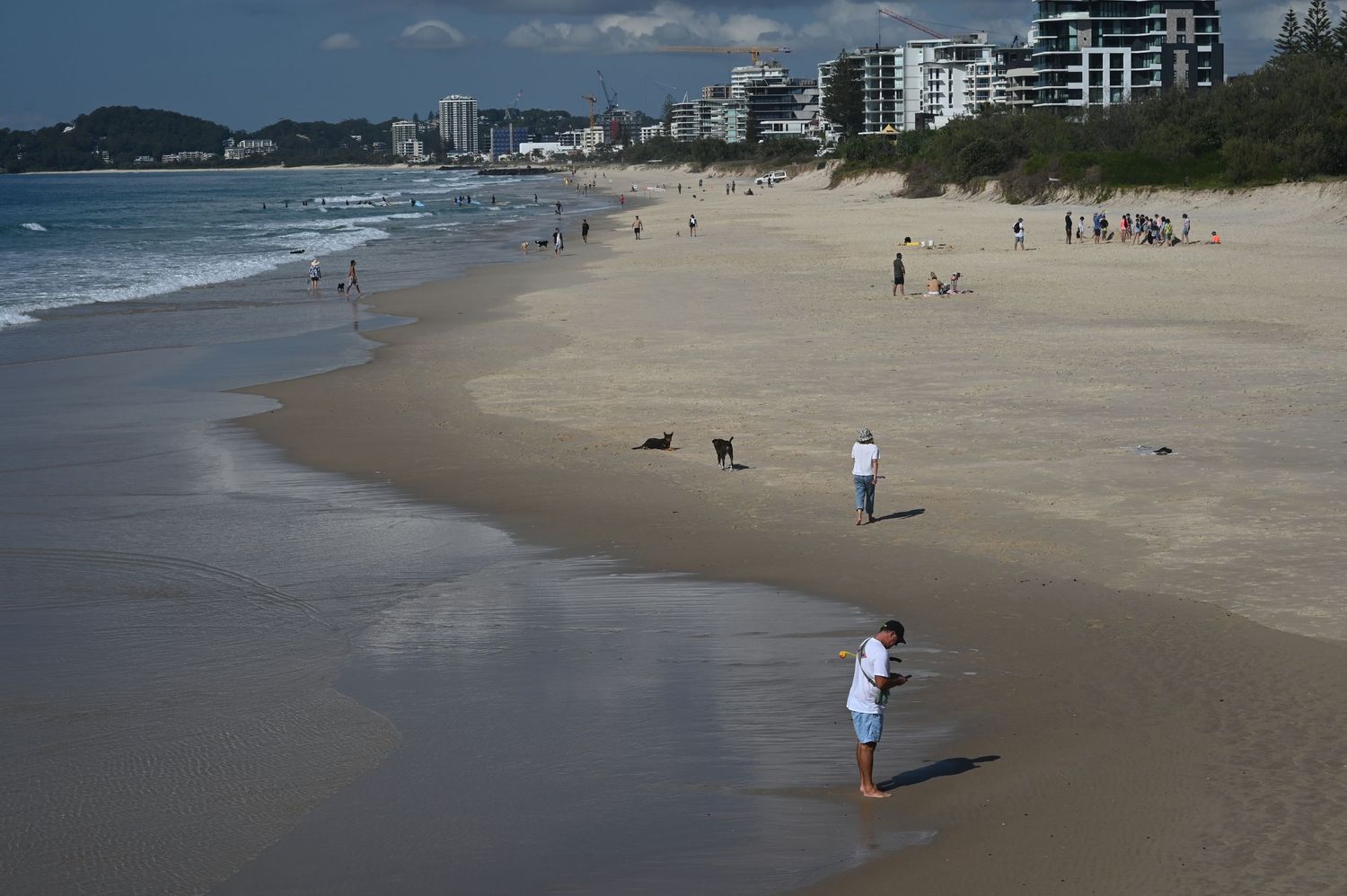 Aerial view of a sandy beach with people walking along the shoreline, with coastal buildings and ocean waves in the background.